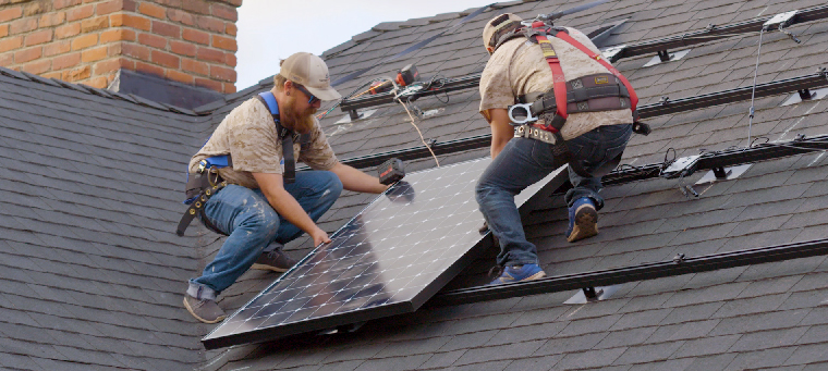 Two solar panel installers on steep, asphalt-shingle roof surrounded by mounting-brackets.