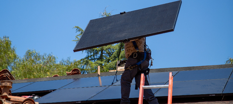 Installer carrying solar panel on roof from a ladder surrounded by new tiles.