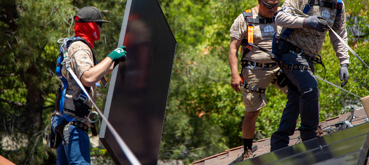 Three installers carrying solar panel on shingle roof surrounded by trees.