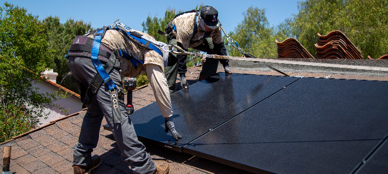 Two installers setting solar panel in place on shingle roof surrounded by trees.