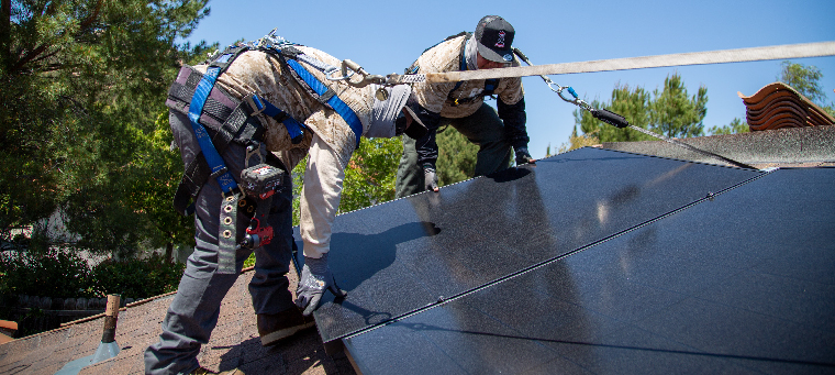Two installers setting solar panel in place on shingle roof.