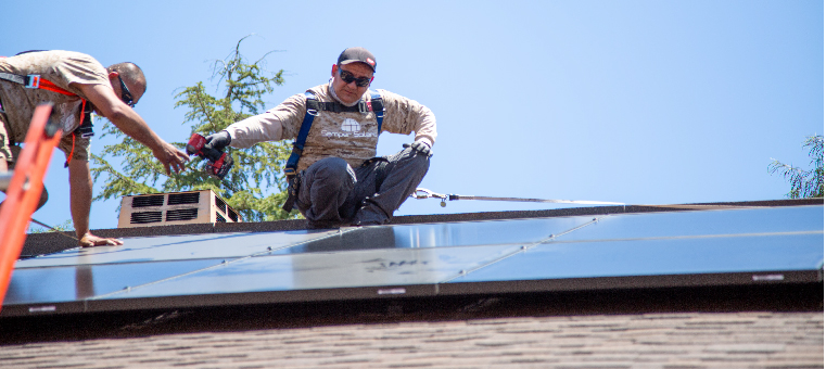 Two installers passing drill to bolt solar panel to shingle roof.