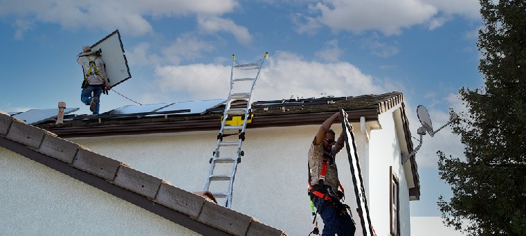 Two installers lifting solar panels onto roof of two story home.