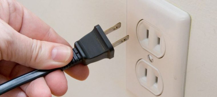 Close-up view of a person’s hand unplugging a black power cord from an outlet strip in a white wall.