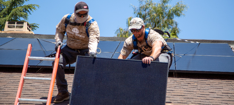 Two installers lifting solar panel onto shingle roof surrounded by trees.